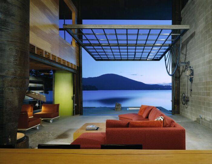 Εσωτερική άποψη του Chicken Point Cabin της Olson Kundig με τον χαρακτηριστικό κινητό γυάλινο τοίχο σε πλήρως ανοιχτή θέση, επιτρέποντας την άμεση ενοποίηση του εσωτερικού χώρου με το φυσικό περιβάλλον και ενισχύοντας τη βιωματική εμπειρία της κατοίκησης.